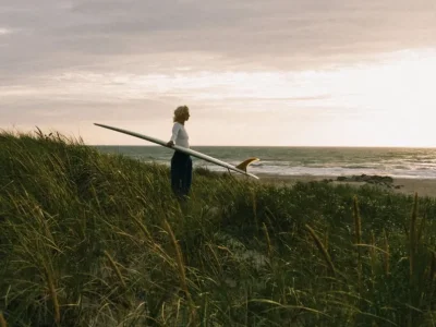 A surfer carrying a board through coastal dunes at sunset, looking out to sea